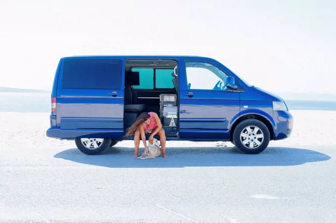 Woman petting dog while sitting on blue motor home at beach against clear sky
