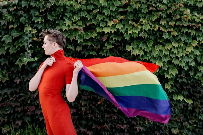 Non-binary person in red dress holding rainbow flag while standing against ivy wall