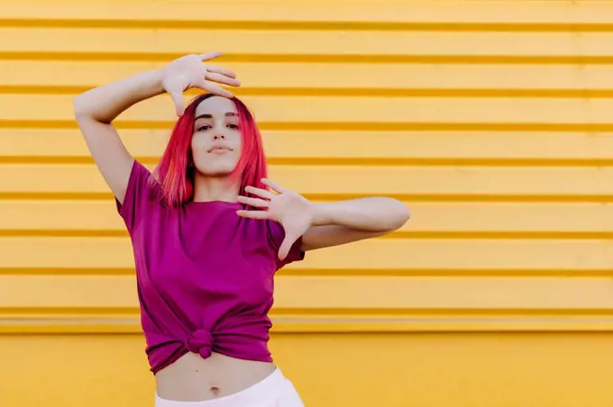 Confident young woman with dyed hair dancing against yellow wall