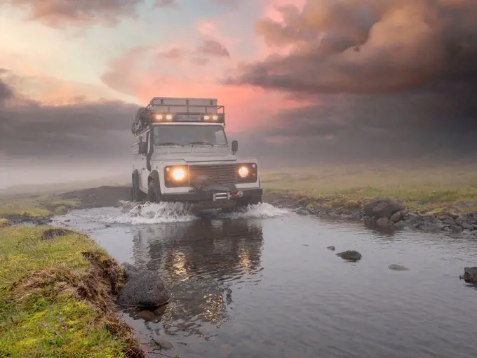 Off-road vehicle splashing water from puddle against cloudy sky at sunset