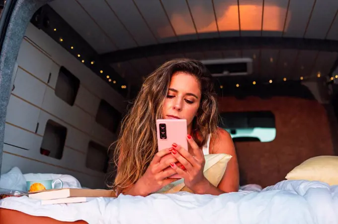 Beautiful woman using mobile phone while lying in camper van during sunset at beach