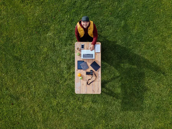 Aerial view of man sitting at coffee table set on green lawn and smiling up at camera