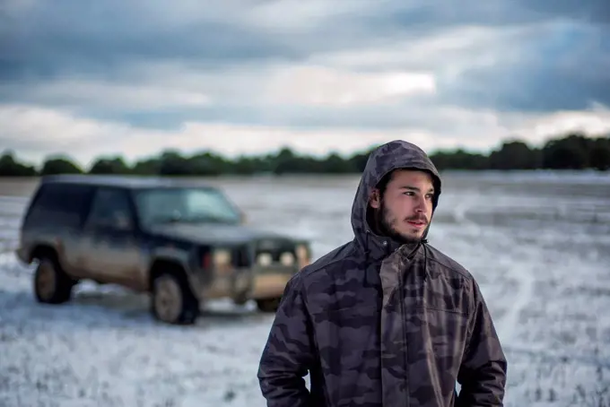 Young man in hooded jacket standing against car at agricultural field