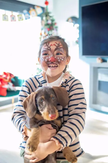 Smiling girl with painted face holding dog at home during Christmas