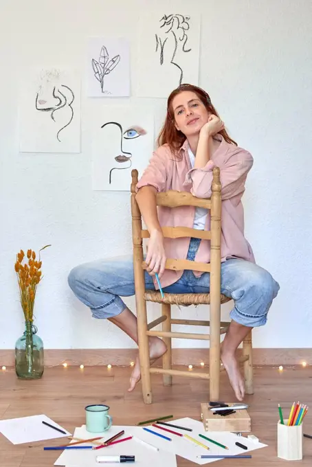 Smiling female artist with hand on chin at chair against wall in living room