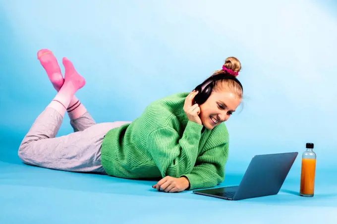 Smiling woman with headphones looking at laptop while lying on front against blue background