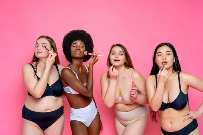 Multi-ethnic group of female models in lingerie applying make-up while standing against pink background