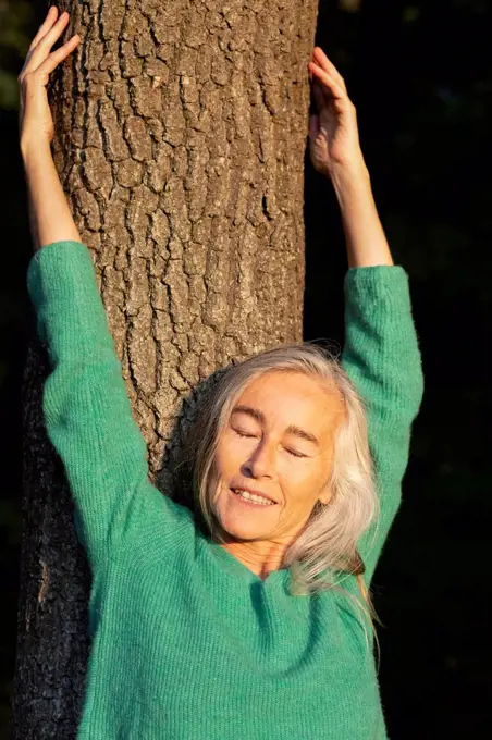 Mature woman with eyes closed against tree trunk at park during autumn
