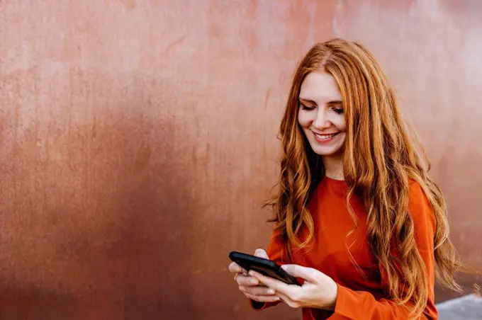 Redhead young woman smiling while text messaging through mobile phone against wall