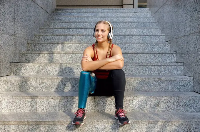 Smiling sportswoman with prosthetic leg wearing headphones sitting with arms crossed on staircase