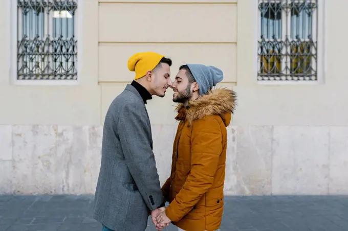 Gay men wearing jacket rubbing noses while holding hands standing against wall