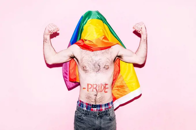 Covered face of gay man with rainbow flag flexing biceps standing against pink background