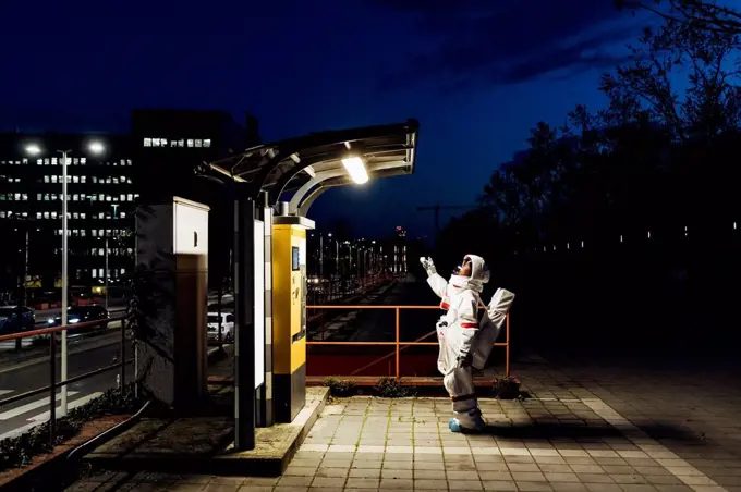 Young female astronaut standing near illuminated telephone booth in city during night