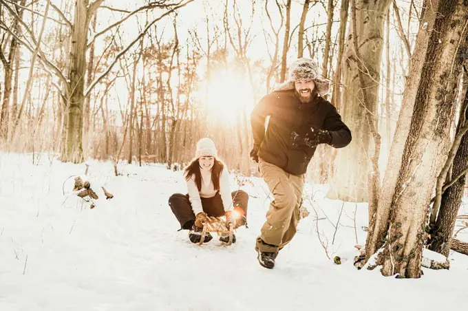 Playful man pulling woman sitting on sled in forest