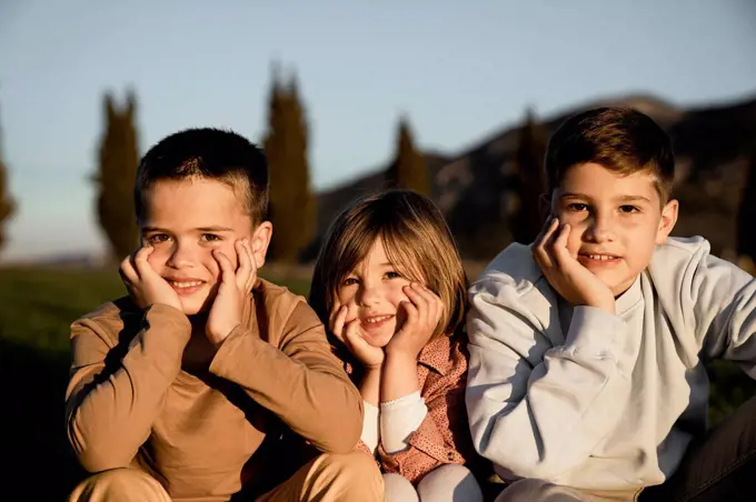 Smiling brothers and sister sitting with hand on chin during sunny day