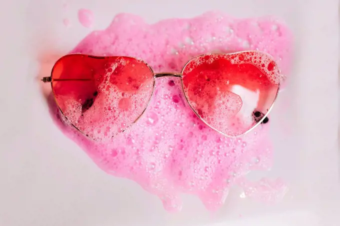 Red heart shape sunglasses on pink soap sud over white background