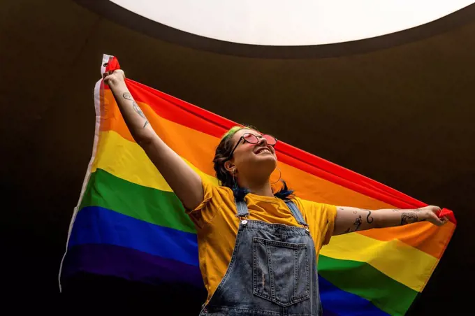 Smiling woman in bib overalls looking up while holding rainbow flag
