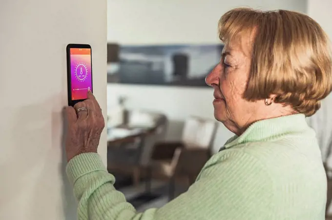 Senior woman using smart thermostat on wall at home