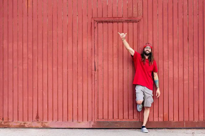 Smiling hipster man making shaka sign while leaning on brown wall
