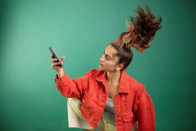 Woman using mobile phone while tossing hair against green background