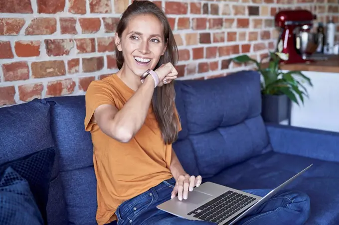 Woman with laptop talking through smart watch while sitting on sofa at home
