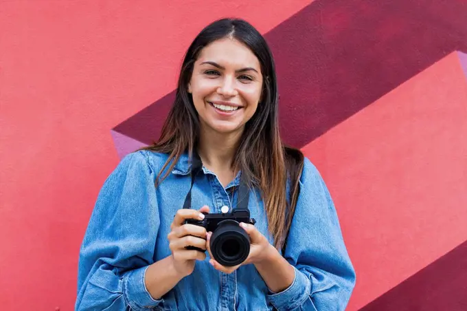 Smiling female freelance worker with camera standing in front of wall