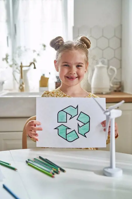 Smiling girl holding recycling symbol drawing on paper at table