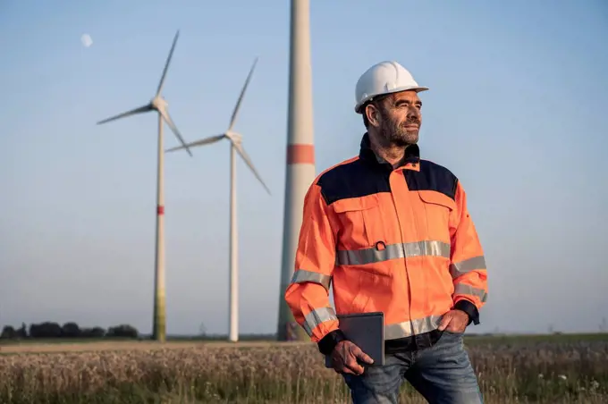 Male engineer holding digital tablet while standing in front of wind turbines