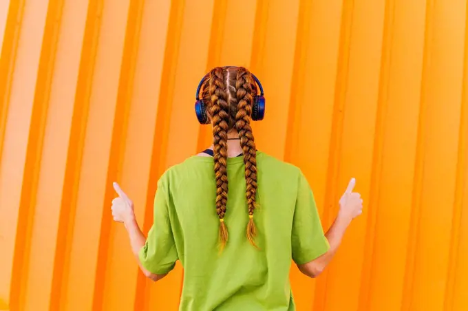 Woman with braided hair gesturing thumbs up while listening music through wireless headphones in front of orange wall