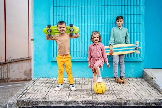 Children standing with skateboards and sports ball on footpath