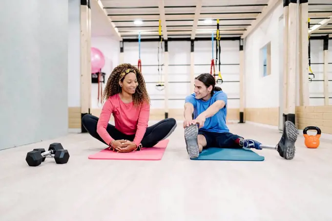 Smiling woman doing stretching exercise with disabled friend at gym