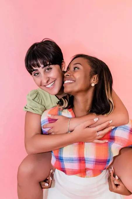 Woman piggybacking lesbian friend against pink background