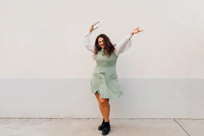 Cheerful plus size woman with smart phone dancing against wall