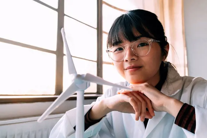Smiling young scientist sitting near wind turbine model in laboratory