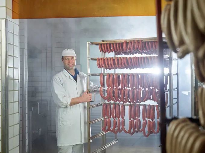 Smiling butcher standing by arranged sausages in smokehouse