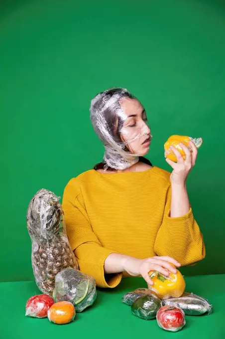 Woman with face and food covered in plastic foil sitting against green background