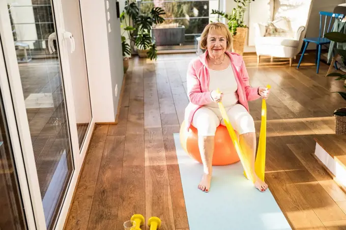 Smiling senior woman sitting on fitness ball exercise with resistance band at home