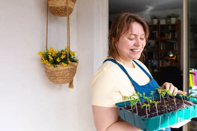 Happy woman with tray of tomato seedlings standing on balcony