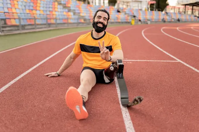 Smiling man with prosthetic leg sitting on running track showing peace sign