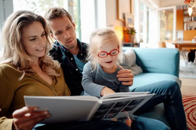 Father and mother looking at cute daughter reading book in living room at home