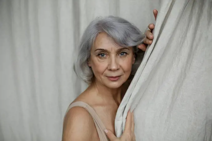 Senior woman in gray hair by curtain at home