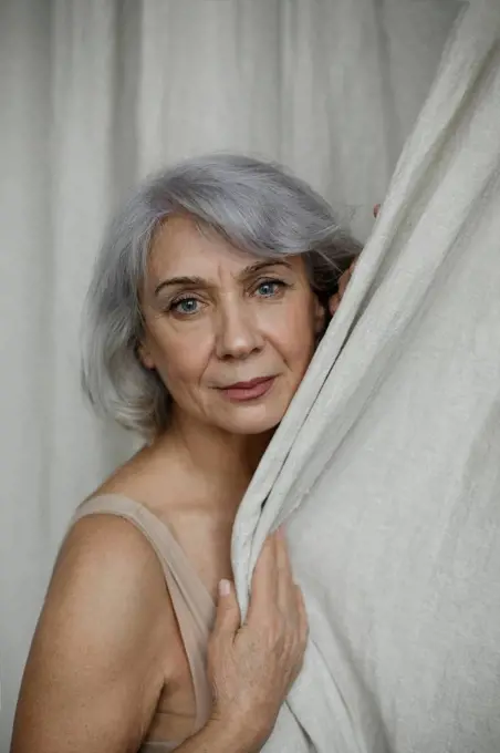 Senior gray haired woman by curtain at home