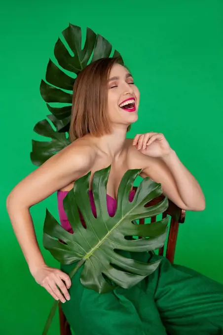 Happy woman sitting on chair with monstera leaf against green background