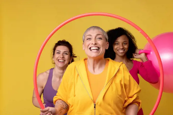 Happy mature woman holding plastic hoop standing with friends against yellow background