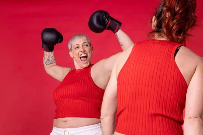 Woman wearing boxing gloves flexing muscles looking at friend against red background
