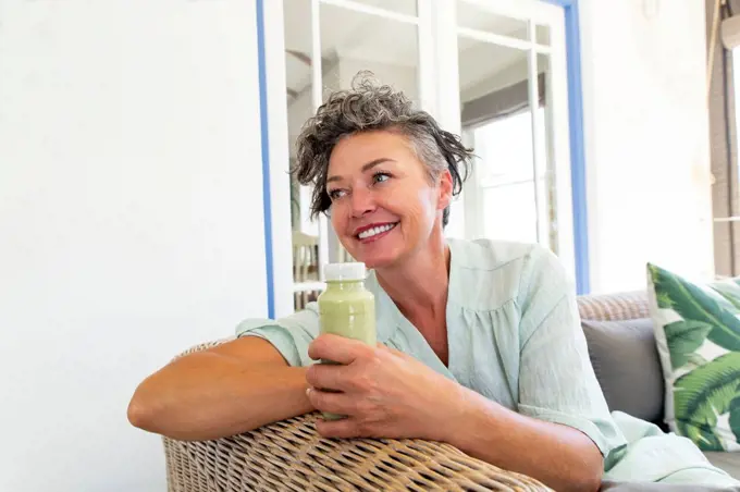 Happy mature woman with bottle of smoothie sitting on sofa at home