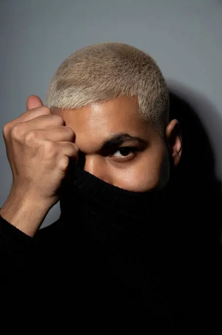 Young man covering face with turtleneck top against gray background