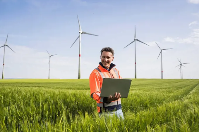 Smiling mature engineer with laptop standing amidst wheat crops on field