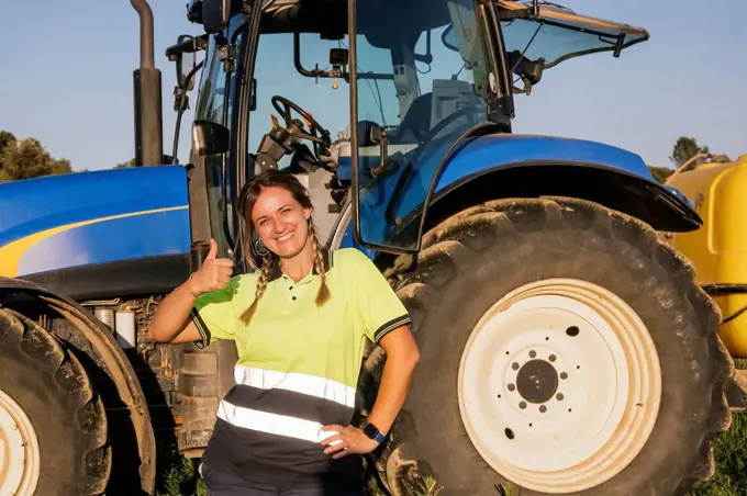 Happy female farmer gesturing thumbs up in front of tractor on sunny day
