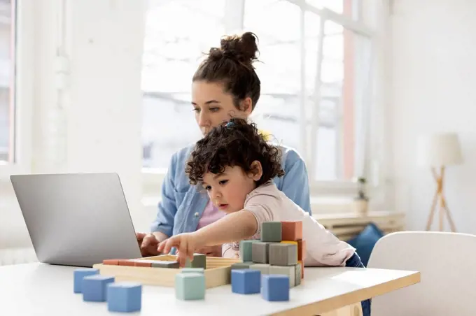 Mother working from home using laptop while daughter is playing with buiilding blocks